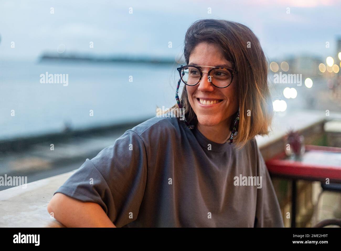 hierba Fundir subasta Mujer joven positiva en ropa casual y anteojos sonriendo y mirando hacia  otro lado mientras se sienta en la cafetería al aire libre cerca del mar en  la noche Cuba Fotografía de