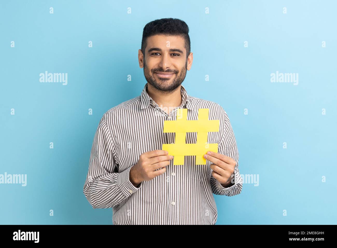 Hashtag viral y blogging exitoso. Hombre de negocios satisfecho con barba sosteniendo gran símbolo de hash amarillo, con camisa de rayas. Estudio de interior disparo aislado sobre fondo azul. Foto de stock