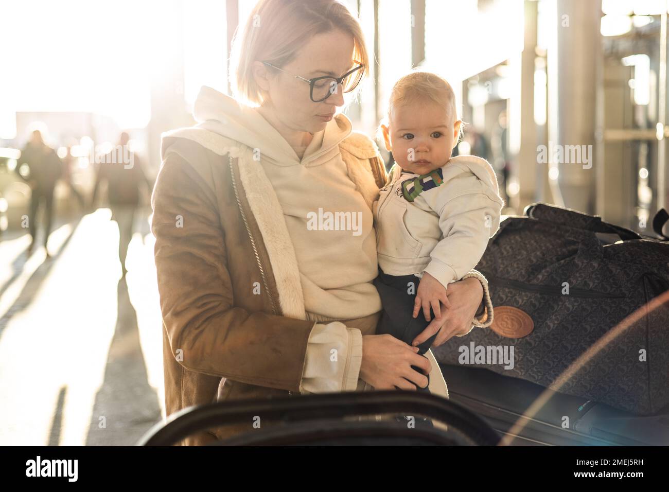 Motherat viaja con su bebé niño, caminando, empujando el cochecito de bebé y el carrito de equipaje frente a la estación terminal del aeropuerto Foto de stock