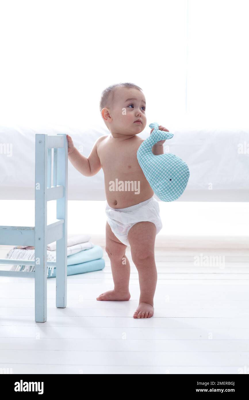 Bebé niño sosteniendo ballena tierna, 11 meses, de pie sosteniendo en la silla de madera blanca Foto de stock