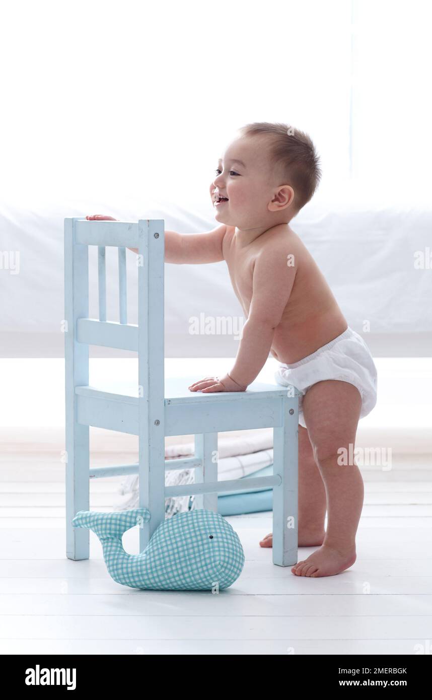 Bebé niño, 11 meses, de pie sosteniendo a la silla de madera blanca Foto de stock