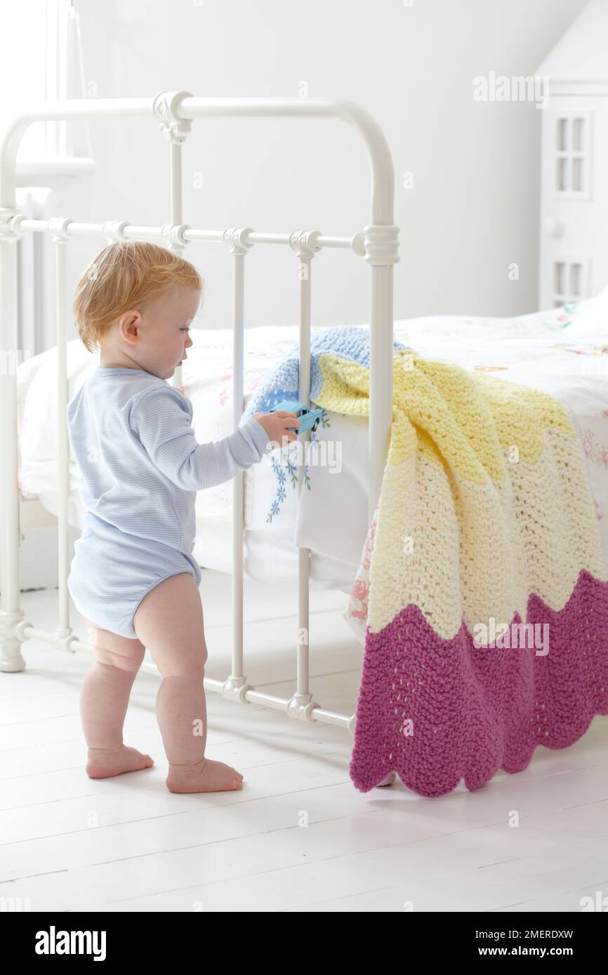 Bebé de pie al pie de la cama jugando con el coche de juguete y la manta tejida, 12 meses Foto de stock