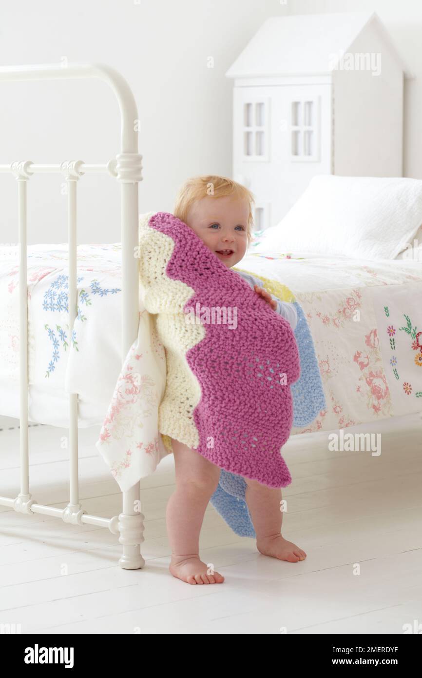 Bebé niño de pie junto a la cama envuelto en manta de punto, 12 meses Foto de stock
