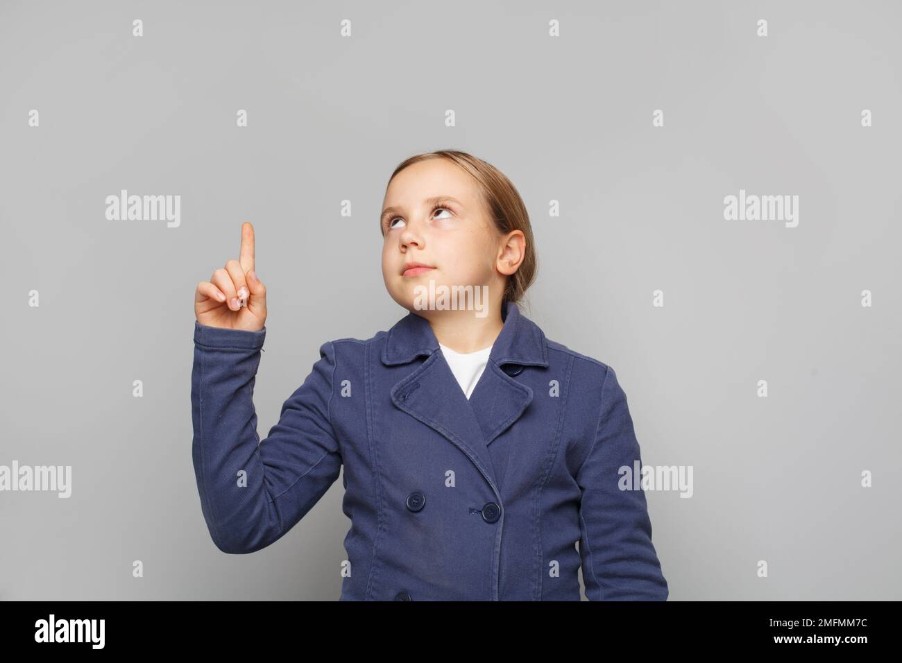 Niña inteligente en uniforme escolar apuntando y mirando hacia arriba contra el fondo gris de la pared del estudio Foto de stock