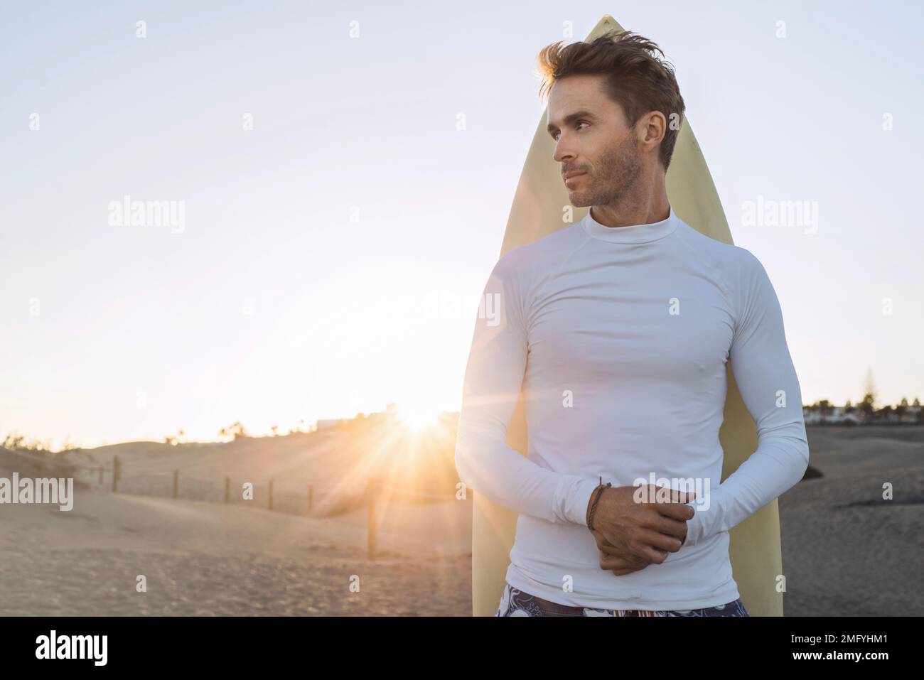Joven chico surfista guapo está de pie junto a su tabla de surf y vestido con camisa blanca, vacía, en blanco de lycra. Maqueta horizontal con destello solar. Foto de stock