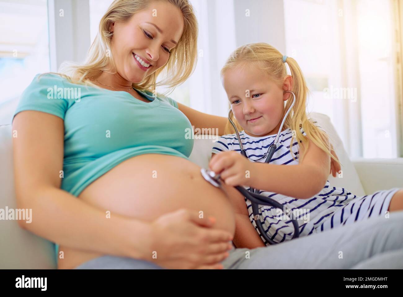 Escuchemos lo que el bebé está diciendo... una niña usando un estetoscopio para escuchar a su madre embarazada del vientre en casa. Foto de stock