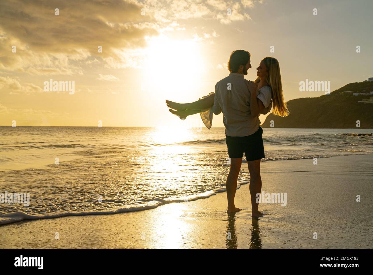 Hombre joven que sostiene a la mujer en la playa durante la puesta del sol mientras se miran en los ojos del otro Foto de stock