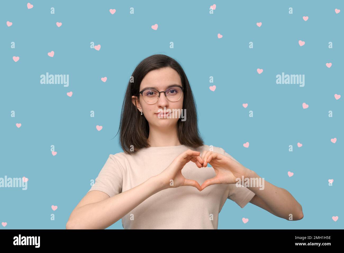 Hermosa chica estudiante en gafas hace una forma de corazón con sus manos, sonríe sobre fondo azul con corazones. Amor, relaciones, día de San Valentín. Foto de stock