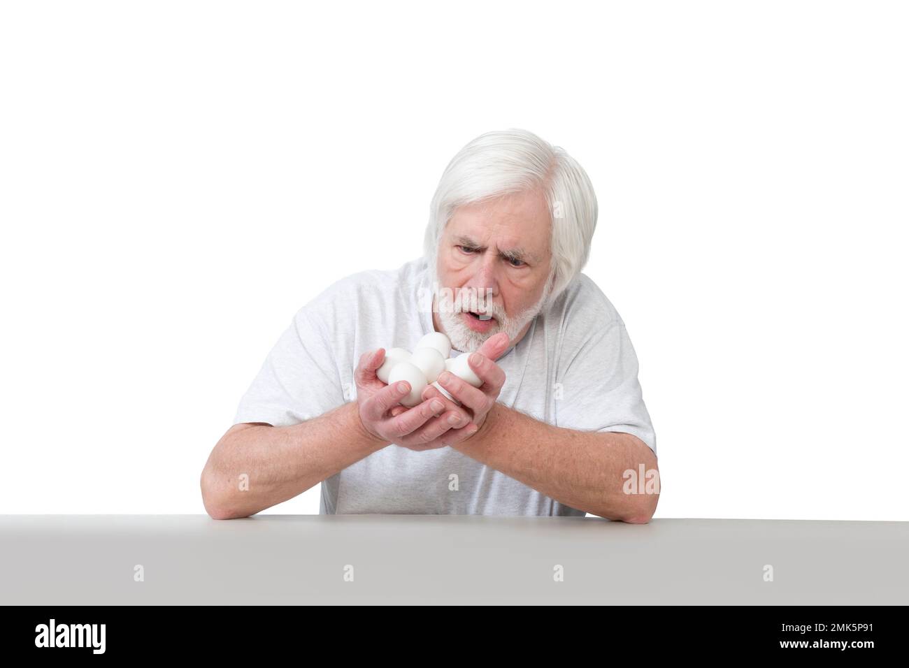 Disparo horizontal de un anciano sentado en una mesa asombrado por su puñado de preciosos huevos blancos de pollo. Aislado en blanco. Un montón de espacio de copia. Foto de stock