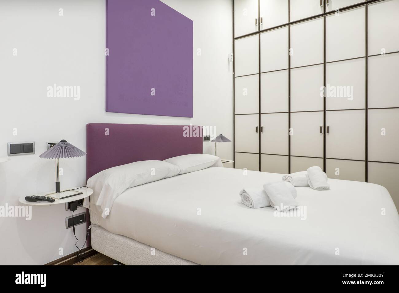 Dormitorio con una cama doble con edredón blanco, almohadas, una cabecera  tapizada púrpura y un armario hecho a medida con puertas blancas y bordes  de madera Fotografía de stock - Alamy