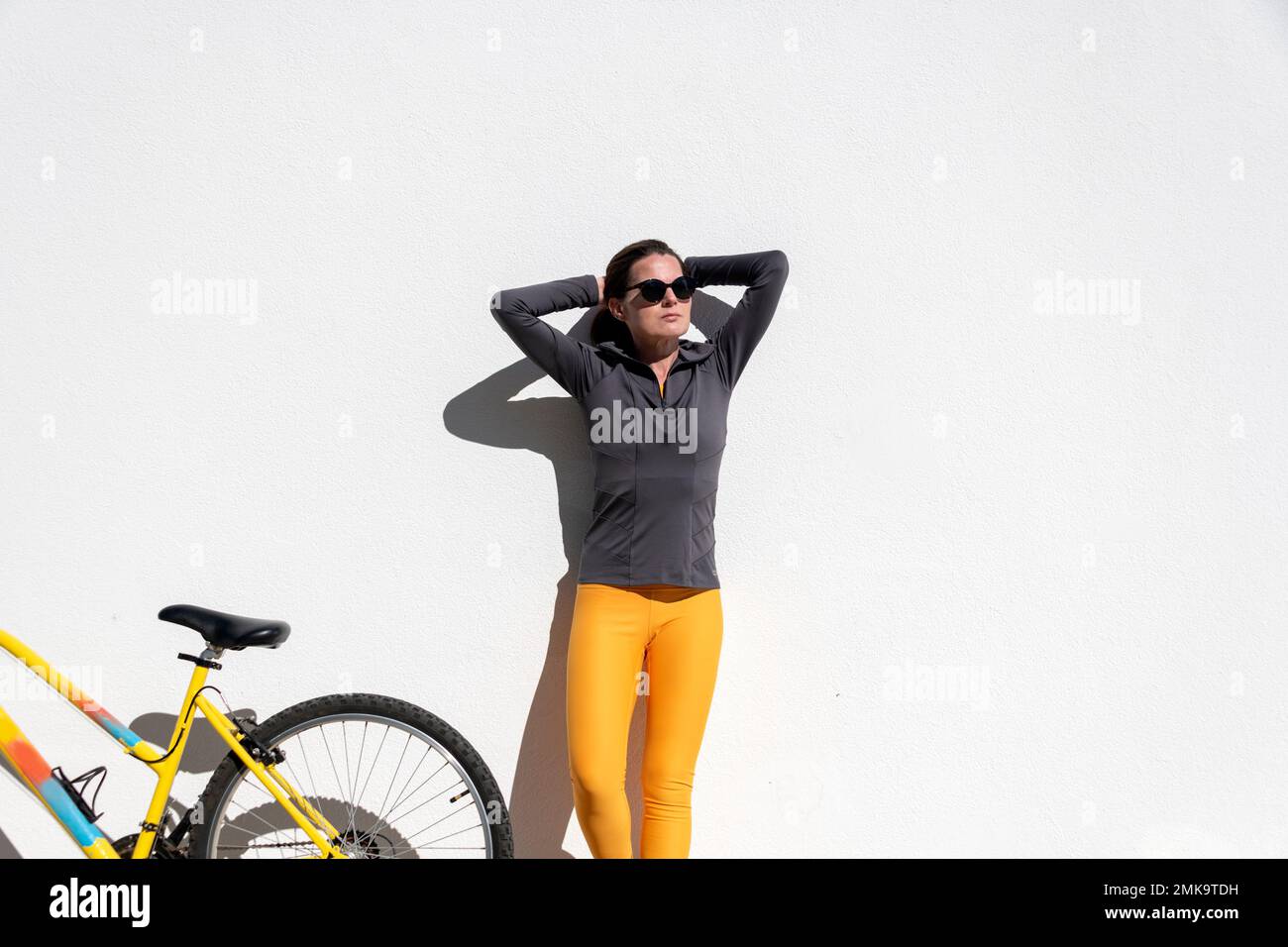 mujer de pie contra una pared con su bicicleta en el sol Foto de stock