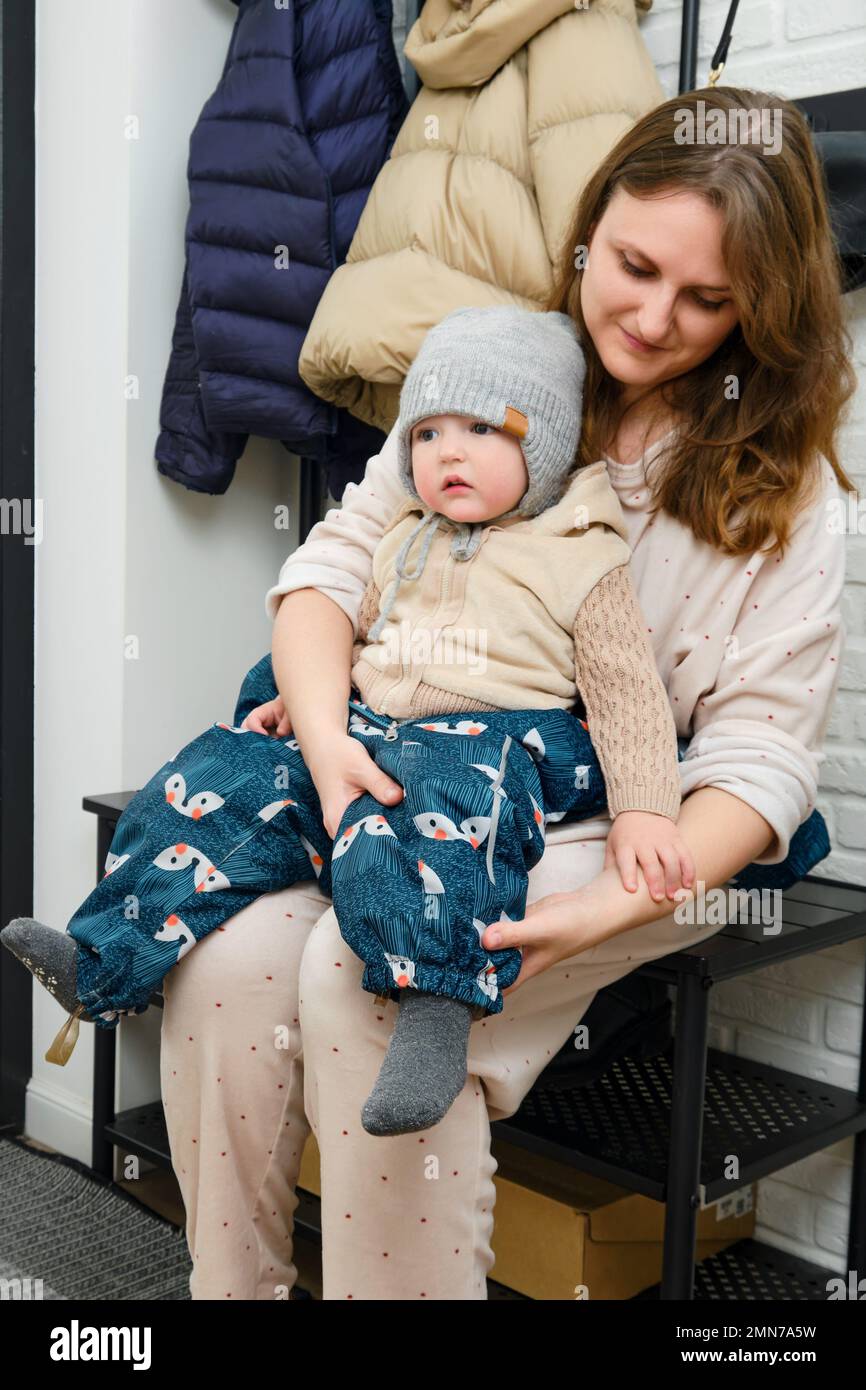 La madre pone un mono azul en el bebé que está sentado en el pasillo de la  casa. Mujer mamá vestir ropa de abrigo sobre el niño para caminar en  invierno en