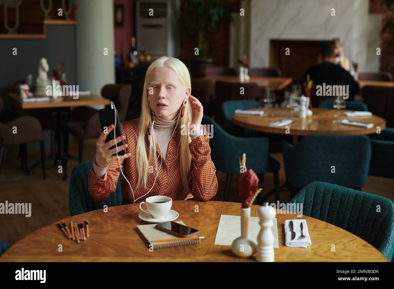 Muchacha bastante albina en ropa casual elegante que se comunica en chat de vídeo con su amiga mientras se sienta junto a la mesa en café moderno en el ocio Foto de stock