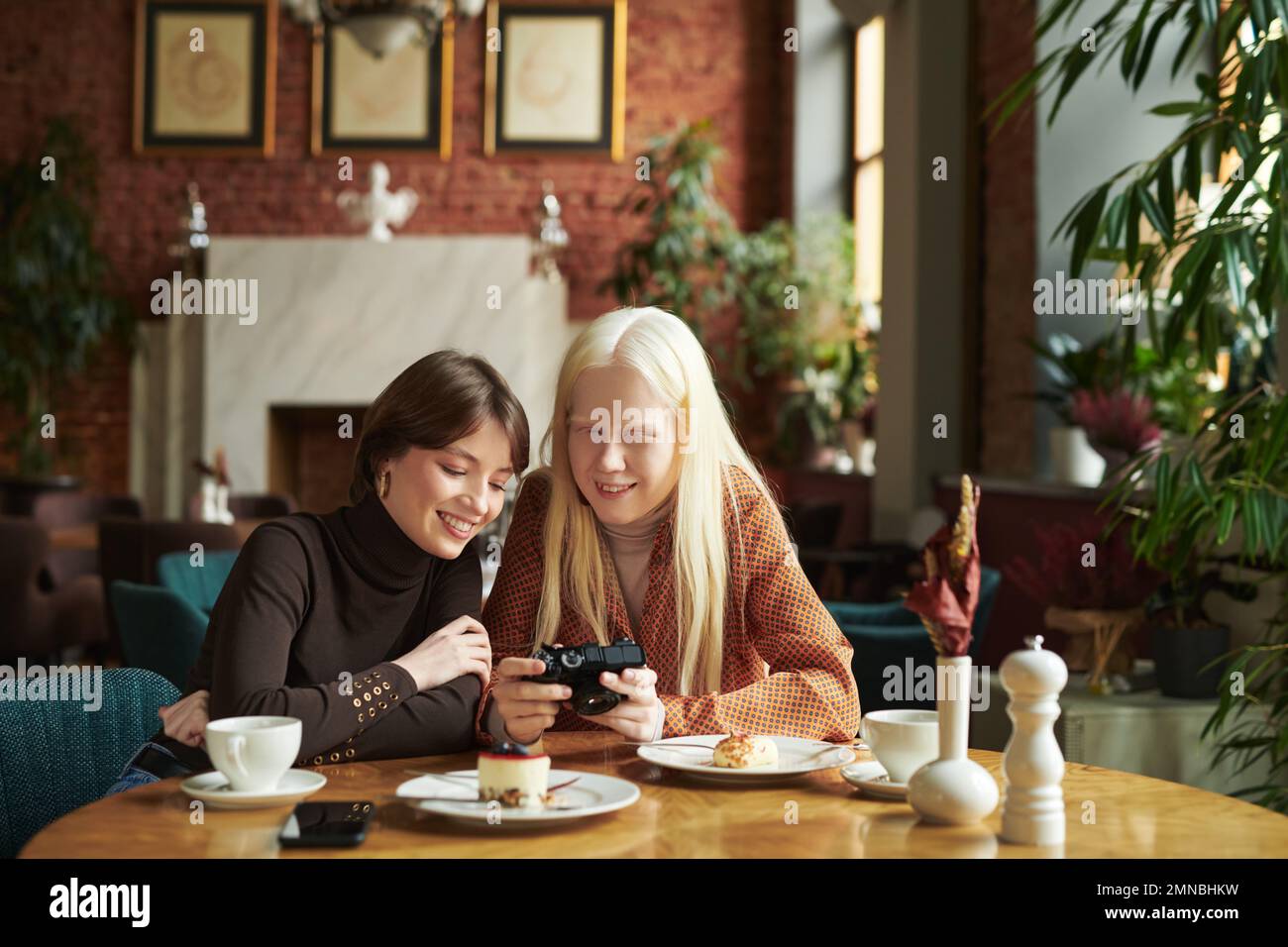 Mujer albina sonriente joven que muestra a su amigo nuevas fotos en cámara fotográfica mientras se sienta junto a la mesa en el café en el almuerzo Foto de stock