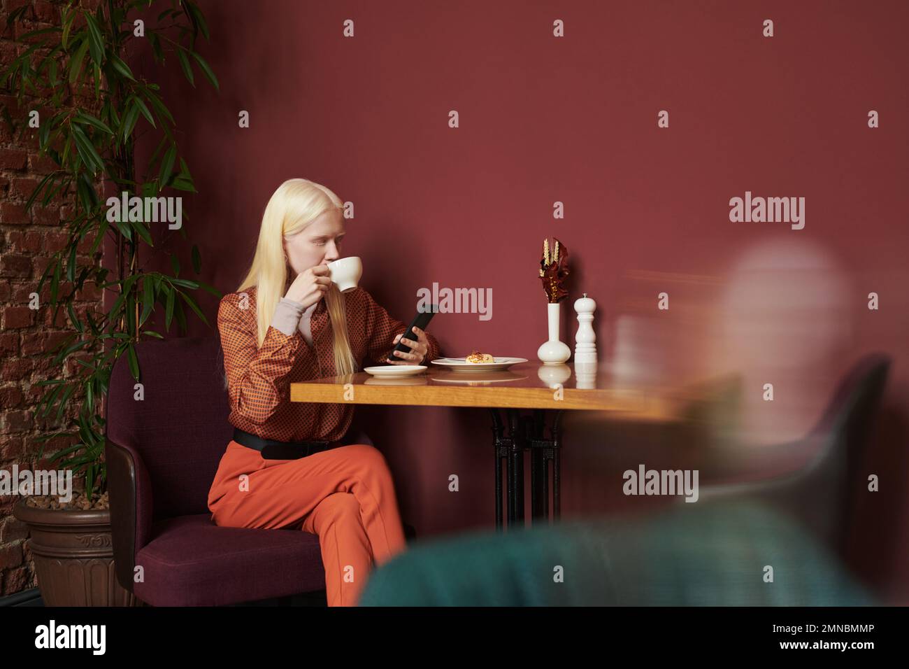 Muchacha albina tranquila en ropa casual elegante tomando café o té con postre y desplazándose en gadget móvil mientras se sienta junto a la mesa en café acogedor Foto de stock