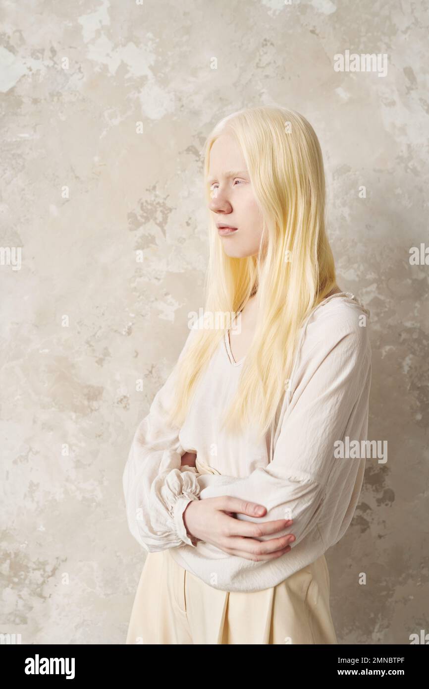 Mujer albina joven en ropa casual de algodón blanco manteniendo sus brazos cruzados por el pecho mientras está de pie junto a la pared de mármol delante de la cámara Foto de stock