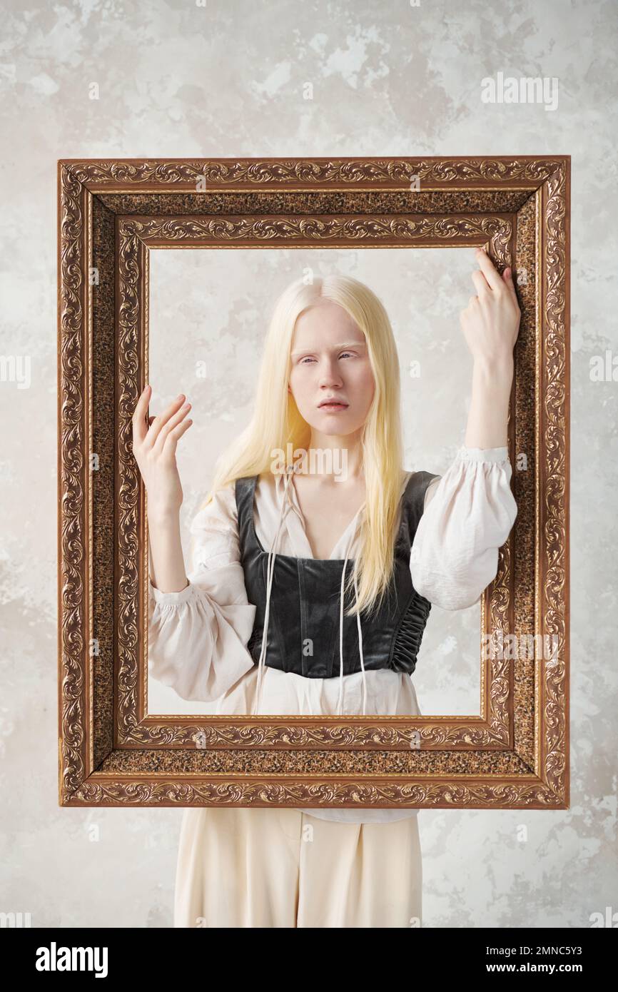 Chica albina linda en atuendo medieval manteniendo las manos en el marco de imagen grande mientras está de pie detrás de él y posando durante la sesión de fotos en el estudio Foto de stock