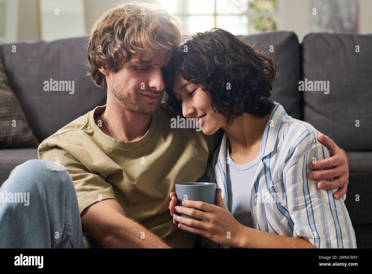 Mujer relajada joven con la taza de café que mantiene la cabeza en el hombro de su marido mientras que ambos disfrutan del ocio o del fin de semana en casa Foto de stock