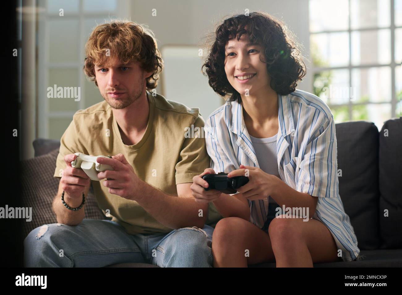 Mujer joven feliz y su novio molesto en atuendo casual sentado en el sofá en la sala de estar en el ocio y jugando al videojuego Foto de stock