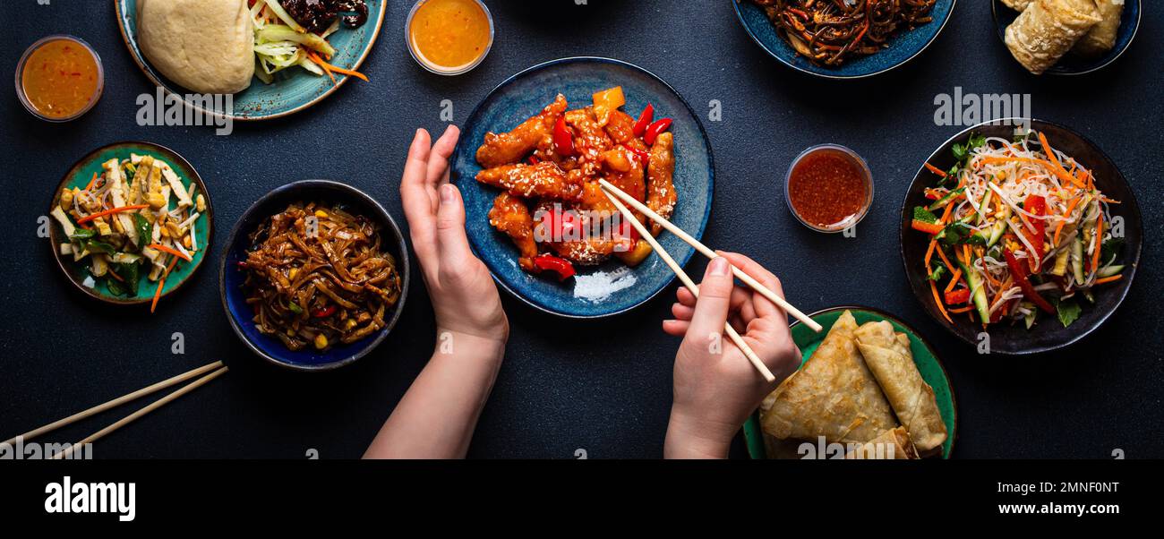 Conjunto de platos chinos en la mesa, manos femeninas sosteniendo palillos: Pollo agridulce, rollitos de primavera fritos, fideos, arroz, bollos al vapor con barbacoa Foto de stock