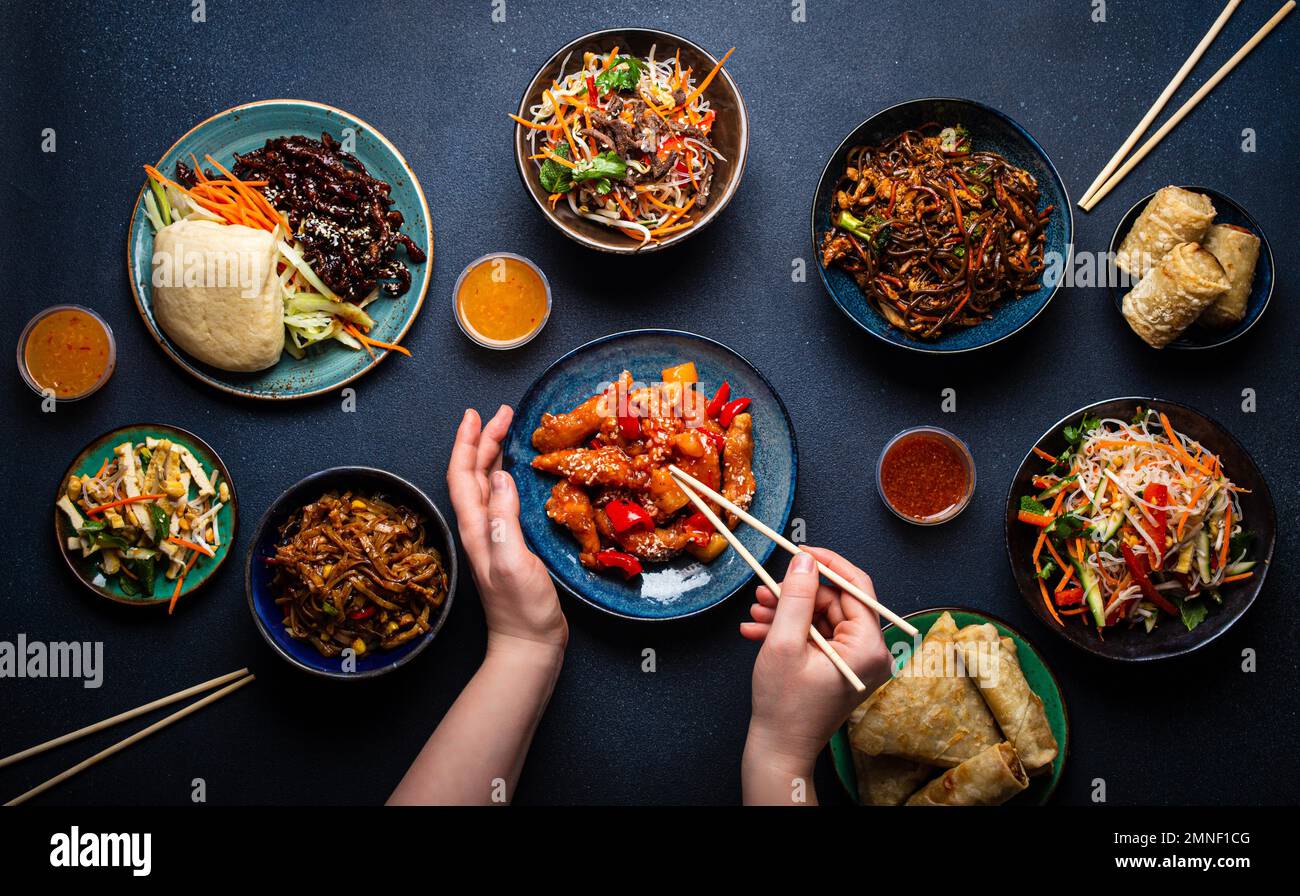 Conjunto de platos chinos en la mesa, manos femeninas sosteniendo palillos: Pollo agridulce, rollitos de primavera fritos, fideos, arroz, bollos al vapor con barbacoa Foto de stock