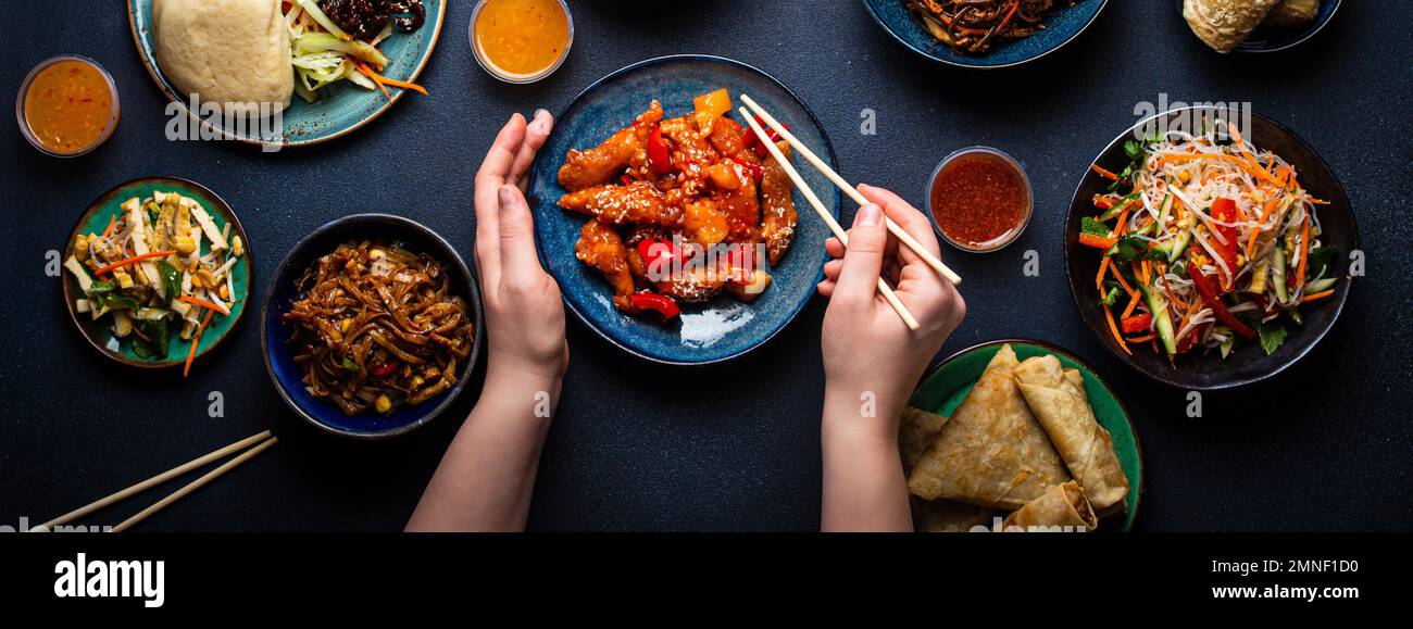 Conjunto de platos chinos en la mesa, manos femeninas sosteniendo palillos: Pollo agridulce, rollitos de primavera fritos, fideos, arroz, bollos al vapor con barbacoa Foto de stock