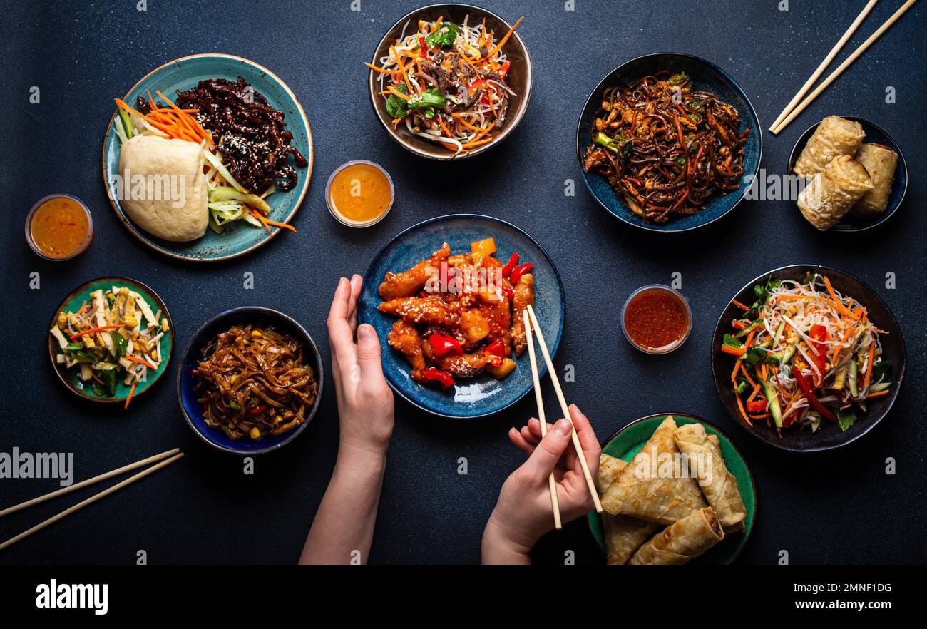 Conjunto de platos chinos en la mesa, manos femeninas sosteniendo palillos: Pollo agridulce, rollitos de primavera fritos, fideos, arroz, bollos al vapor con barbacoa Foto de stock