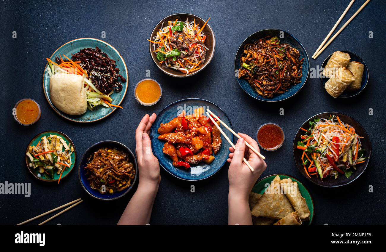 Conjunto de platos chinos en la mesa, manos femeninas sosteniendo palillos: Pollo agridulce, rollitos de primavera fritos, fideos, arroz, bollos al vapor con barbacoa Foto de stock