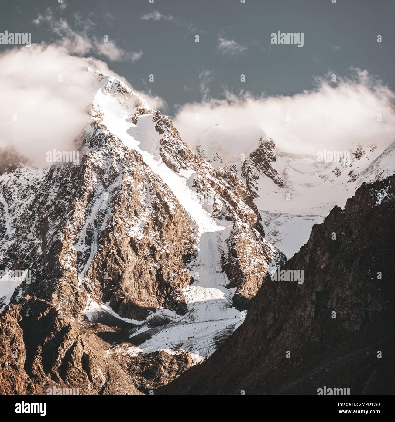 La lengua de un glaciar alpino con nieve desciende de altas montañas de piedra rocosa entre los picos en las nubes en Altai a la sombra durante el día. Foto de stock