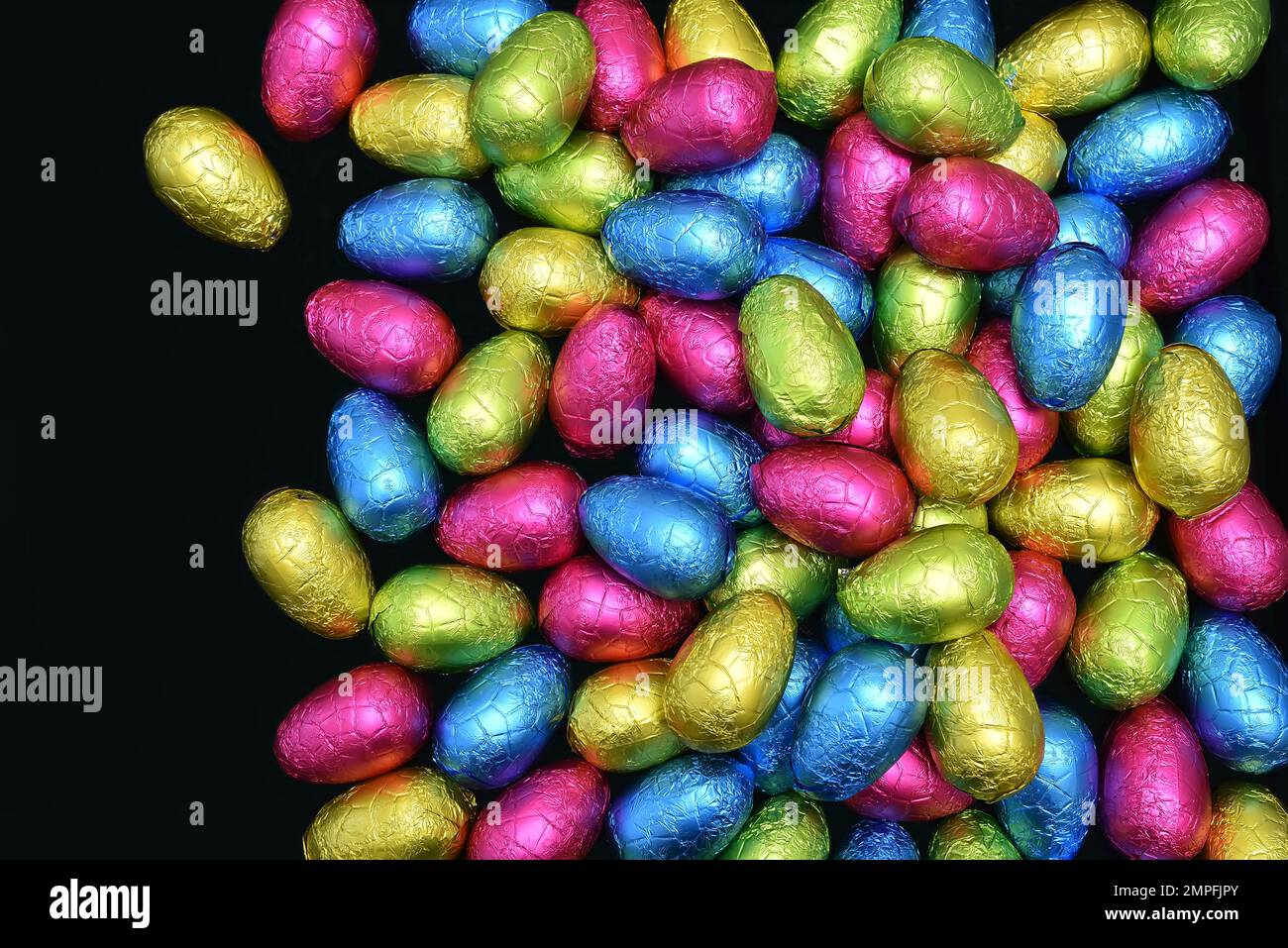 Pila de varios colores y diferentes tamaños de huevos de pascua de chocolate envueltos en papel de aluminio en rosa, azul, amarillo y verde lima sobre un fondo negro. Foto de stock