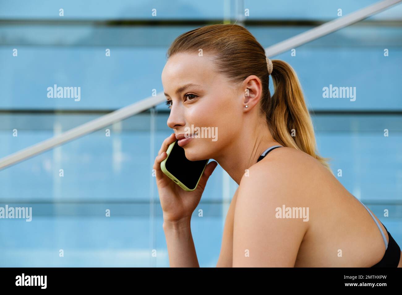 Mujer rubia joven blanca con sujetador deportivo usando teléfono móvil mientras se sienta al aire libre Foto de stock