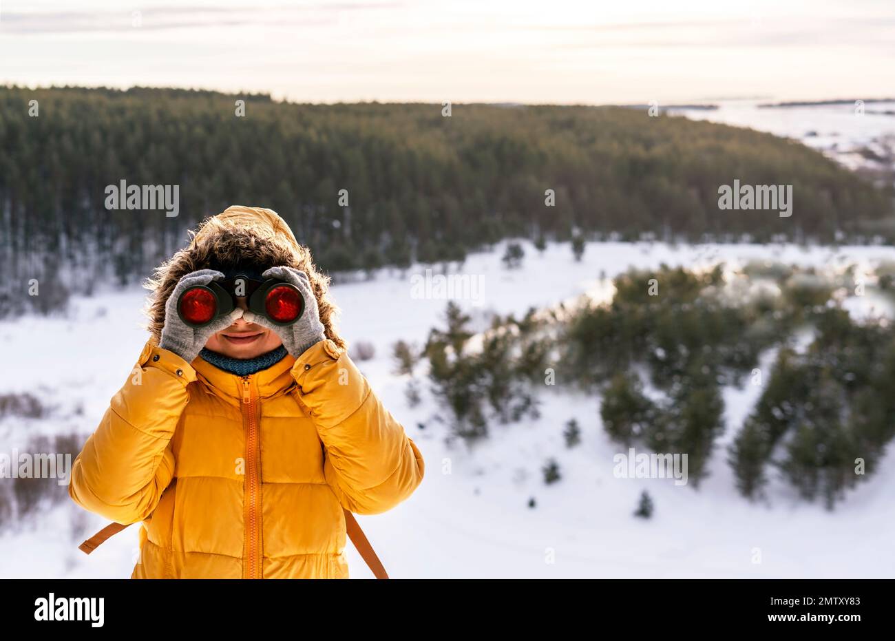 Mujer sonriente joven en amarillo mirando a través de binoculares en aves en río nevado contra bosque de invierno Birdwatching, zoología, ecología Investigación en natu Foto de stock