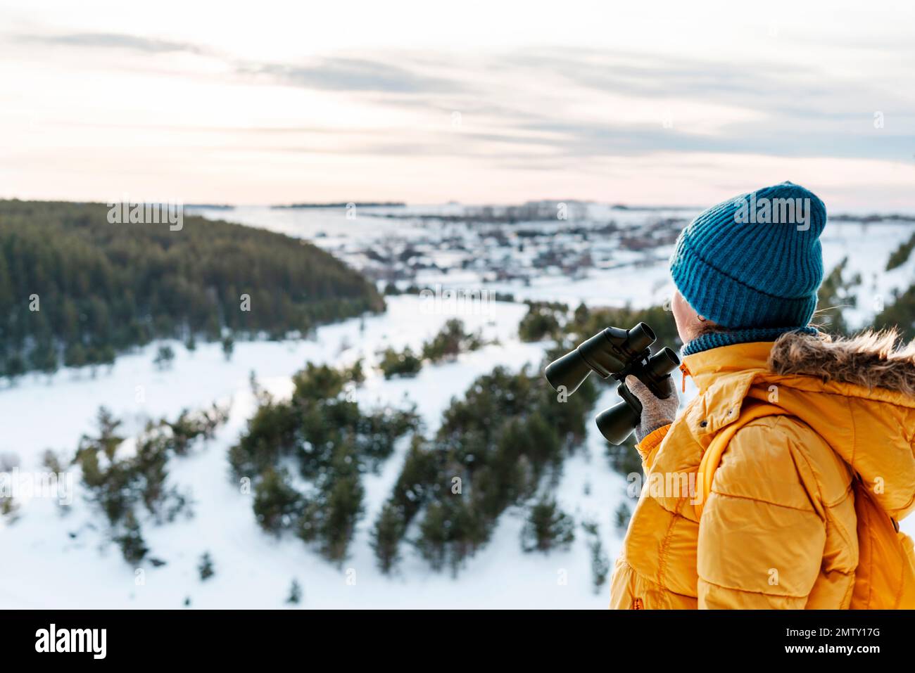 Mujer joven en amarillo con binoculares mirando a los pájaros en la montaña contra el río nevado del invierno y el bosque Birdwatching, investigación de la ecología de la zoología en nat Foto de stock