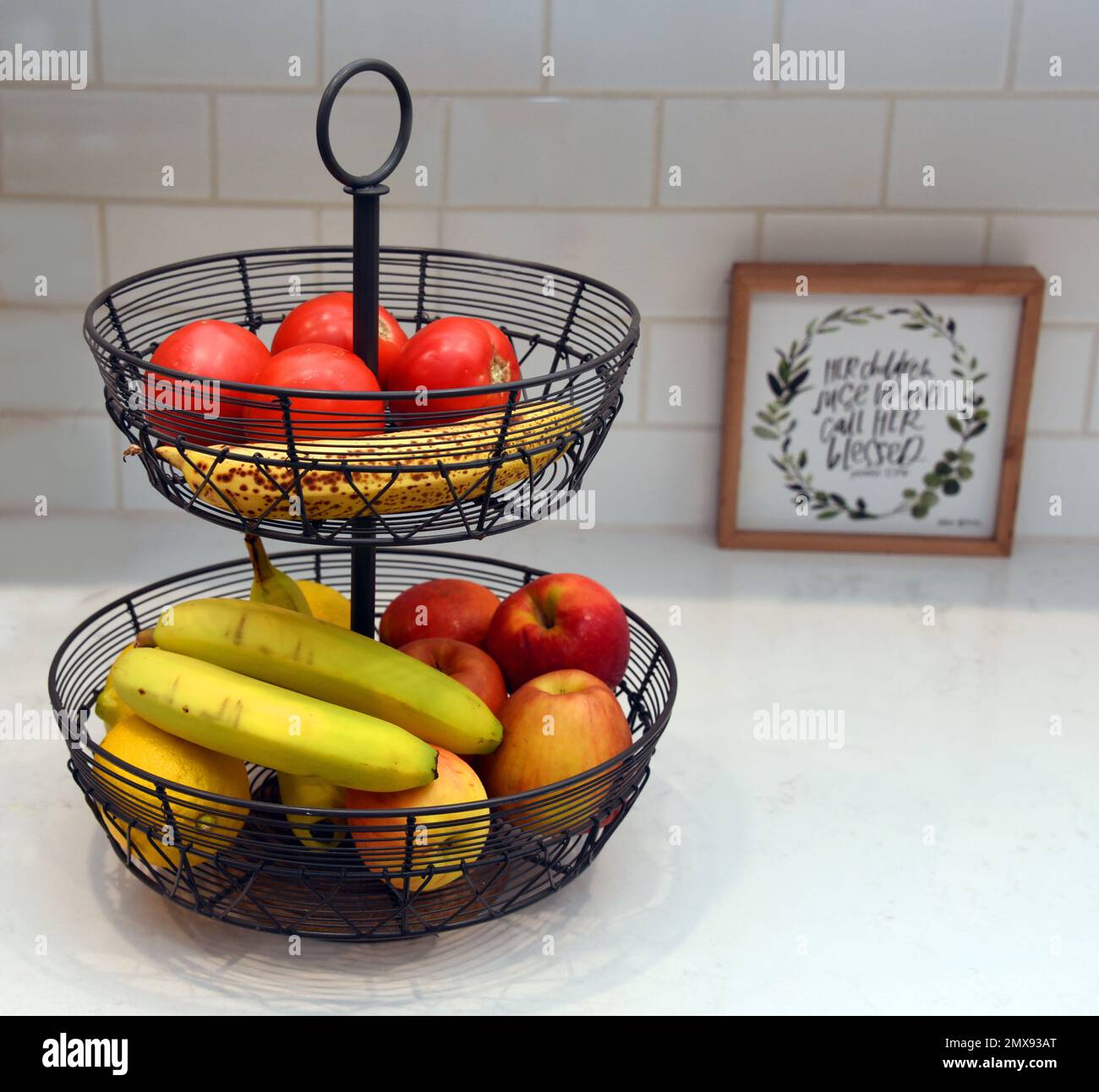 Dos niveles, negro, hierro, cesta de fruta se sienta en una encimera blanca de la cocina. Las frutas incluyen plátanos, tomate, manzanas y limón. Foto de stock