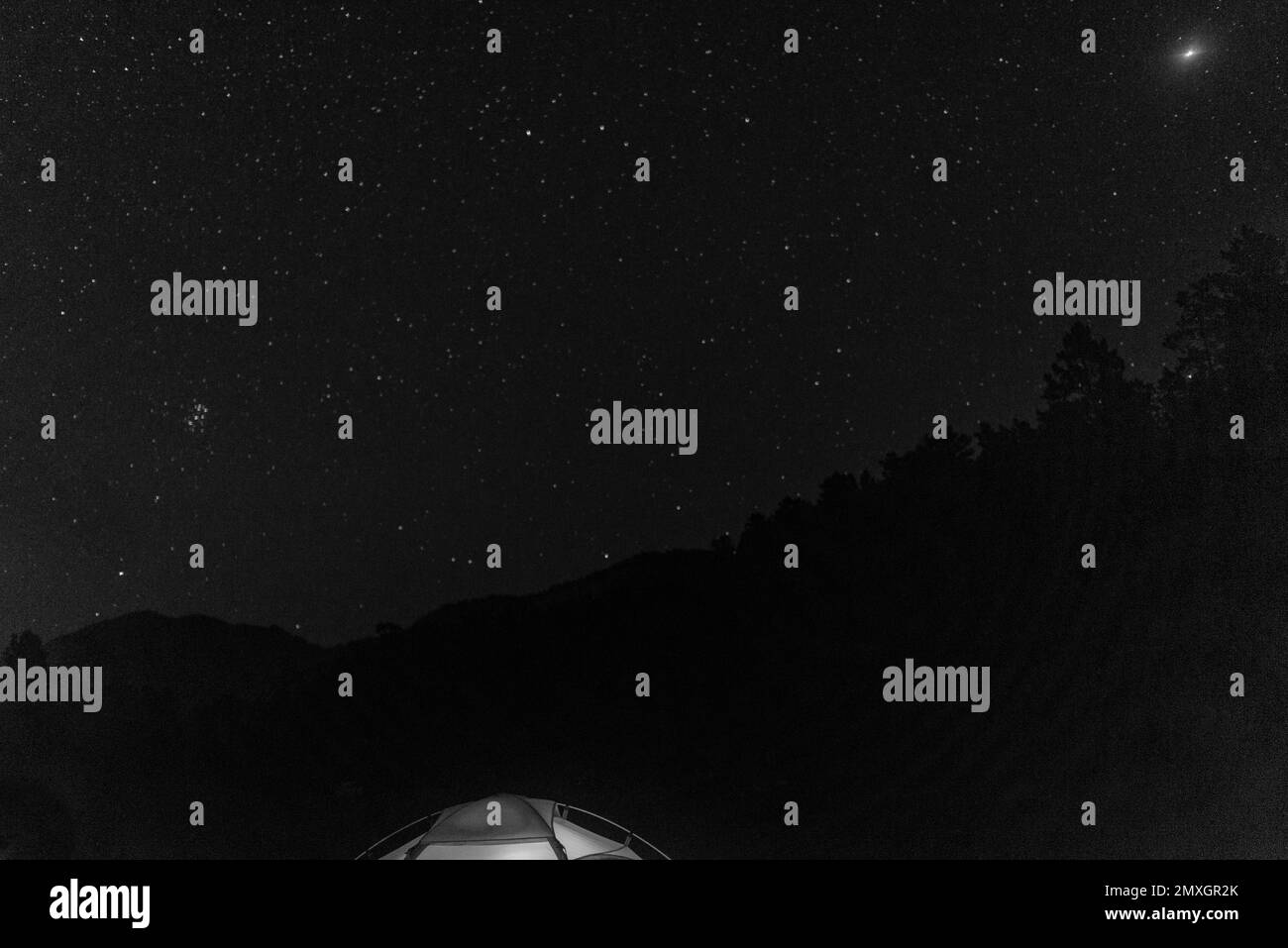 Foto en blanco y negro del borde de una tienda de campaña turística sobre un cielo estrellado en las montañas boscosas por la noche. Foto de stock