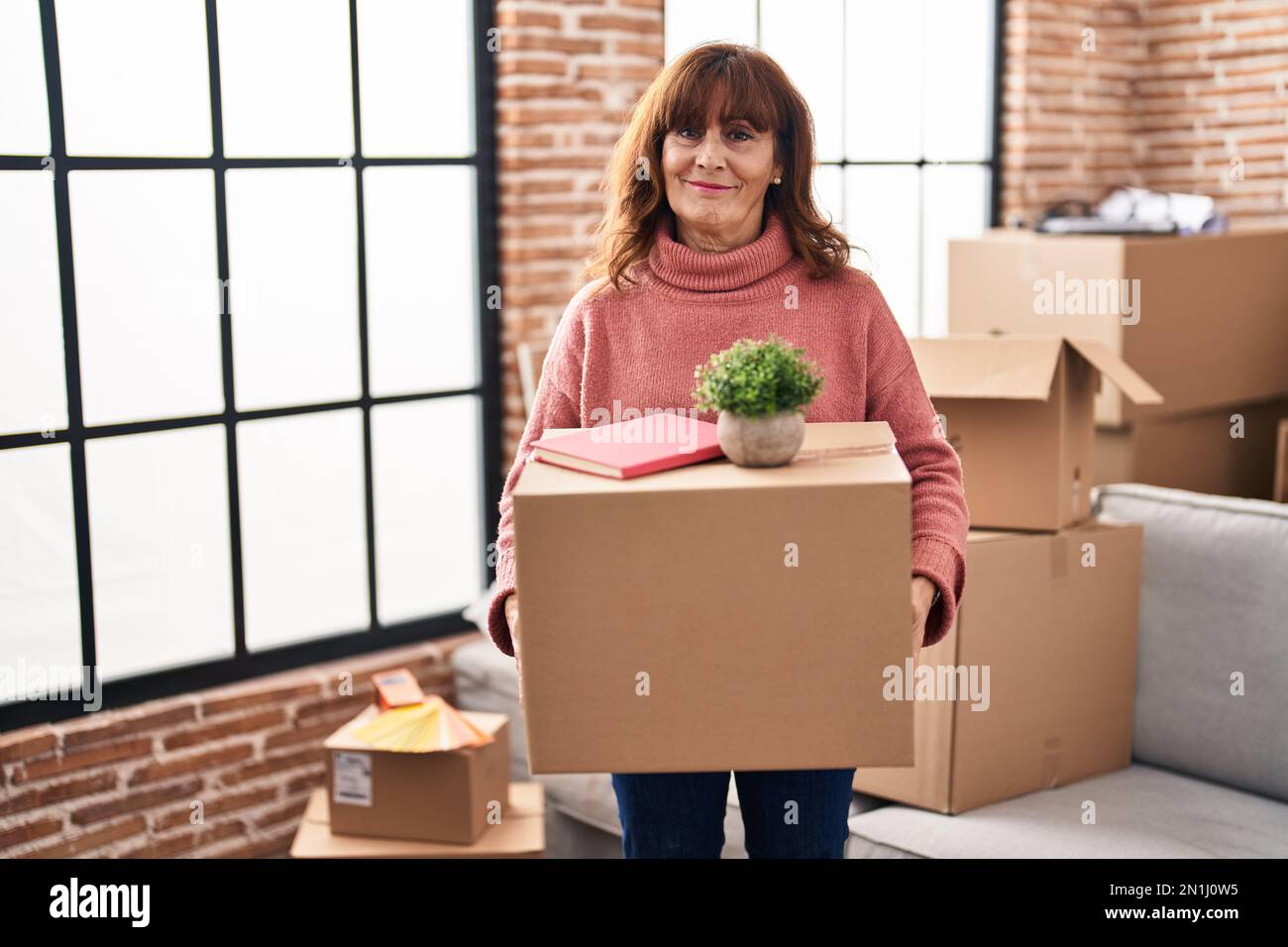 Mujer hispana de edad media que se muda a un nuevo hogar que sostiene la  caja de cartón relajada con expresión seria en la cara. simple y natural  mirando a la cámara