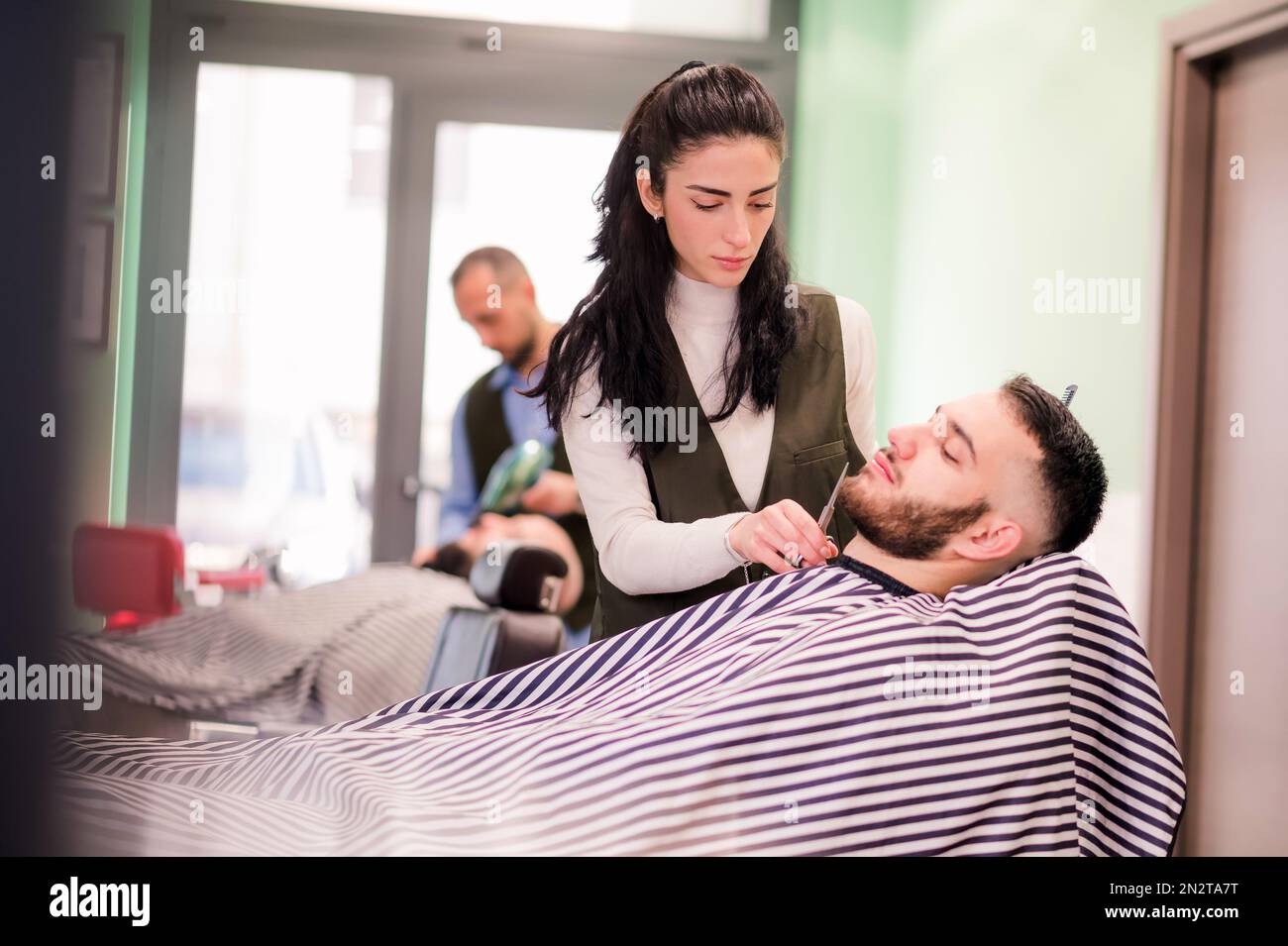 Peluquero femenino profesional concentrado que ajusta la barba con tijeras de cliente masculino en barbería moderna Foto de stock