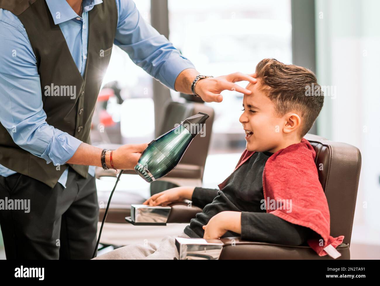 Vista lateral del barbero anónimo de la cosecha en el pelo de los niños de secado uniforme sentado en el sillón en la barbería moderna Foto de stock