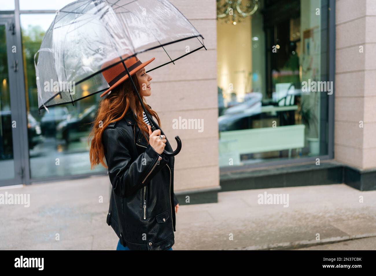 Vista lateral de la joven pelirroja encantadora en elegante sombrero  caminando en la hermosa calle de la ciudad con el paraguas transparente que  disfruta del tiempo lluvioso al aire libre Fotografía de
