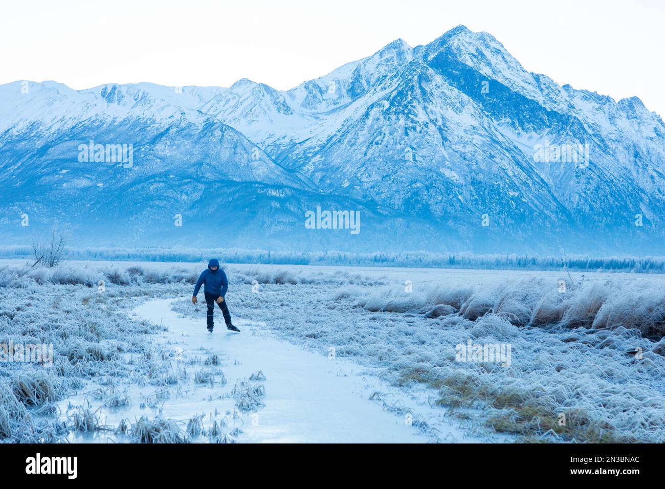 Hombre caucásico patinaje sobre hielo de fondo, blading nórdico, en el sol de invierno en un pequeño arroyo forrado con hierba cubierta de heladas con Pioneer Peak y el chug... Foto de stock