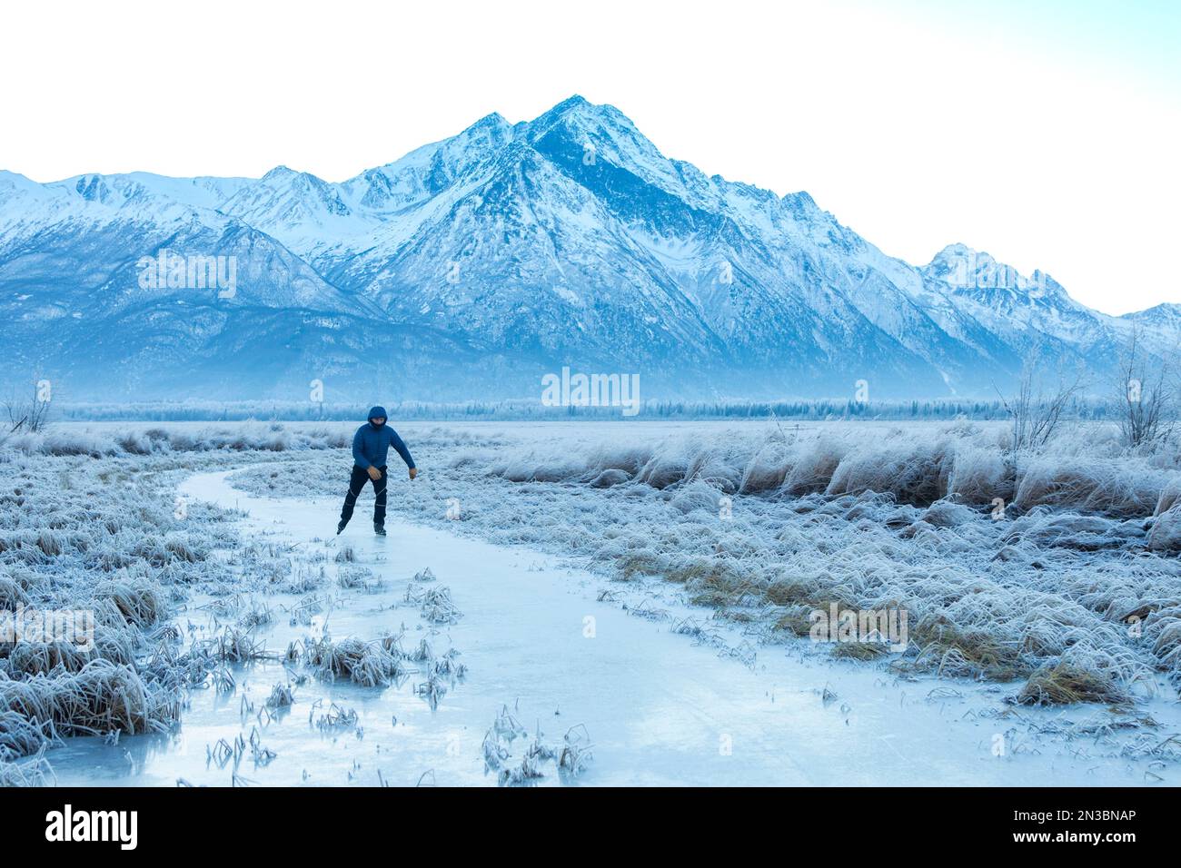 Hombre caucásico patinaje sobre hielo de fondo, blading nórdico, en el sol de invierno en un pequeño arroyo forrado con hierba cubierta de heladas con Pioneer Peak y el chug... Foto de stock