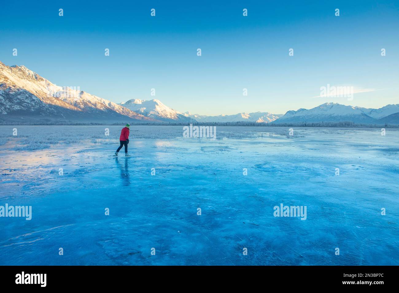 Una mujer caucásica patinaje sobre hielo de fondo, blading nórdico en Gull Lake en Palmer, en un día de invierno con el sol iluminando las montañas Chugach en... Foto de stock