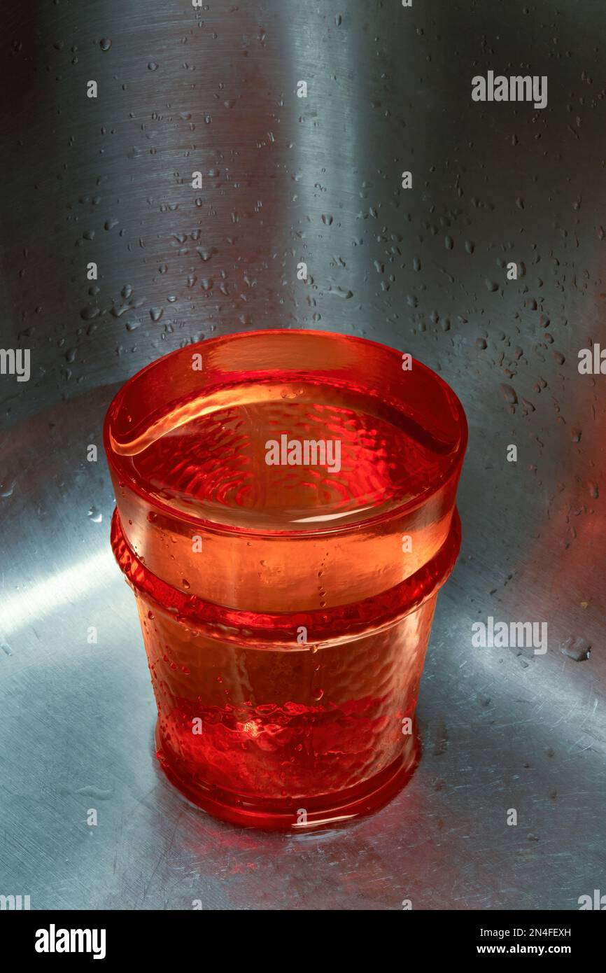 Un vaso rojo lleno de agua sentado en la esquina del fregadero de acero inoxidable con espacio de copia inversa arriba. Foto de stock