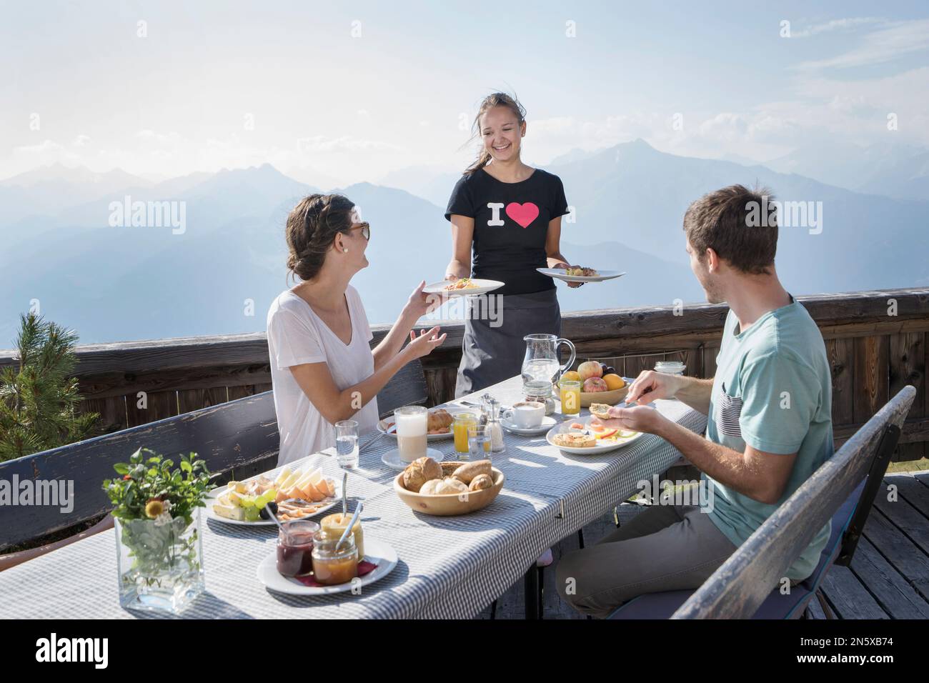 Mujer joven que sirve el desayuno a la pareja en la terraza, Zillertal, Tirol, Austria Foto de stock