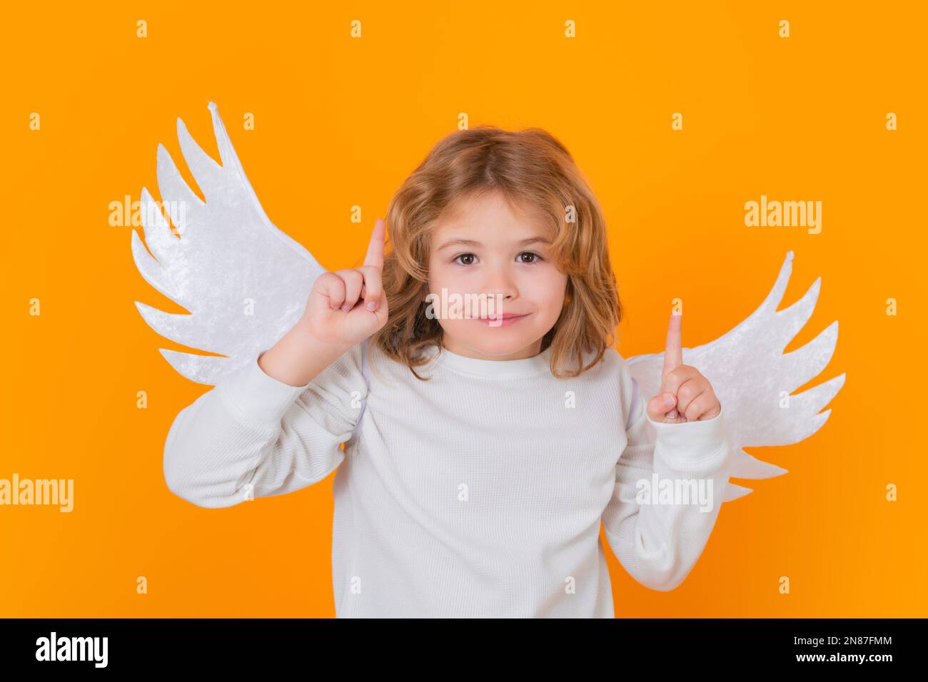 Ángel niño apuntando hacia arriba, gesto de punto, idea. Día de San Valentín. Niño lindo rubio con alas de ángel en un fondo de estudio amarillo. Niño ángel feliz. Foto de stock