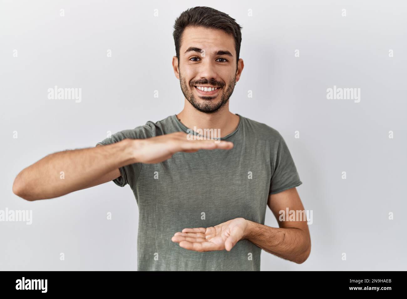 Hombre hispano joven con la barba que lleva la camiseta casual sobre fondo  blanco gesticulando con las manos que muestran el signo grande y grande del  tamaño, símbolo de la medida. sonriente