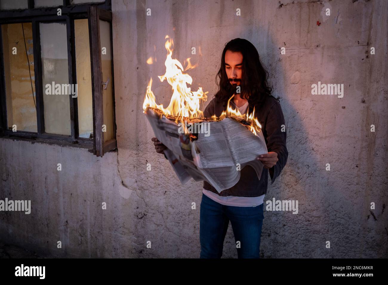 Hombre guapo con el pelo largo leyendo un periódico en llamas en un espacio oscuro en ruinas. Foto de stock