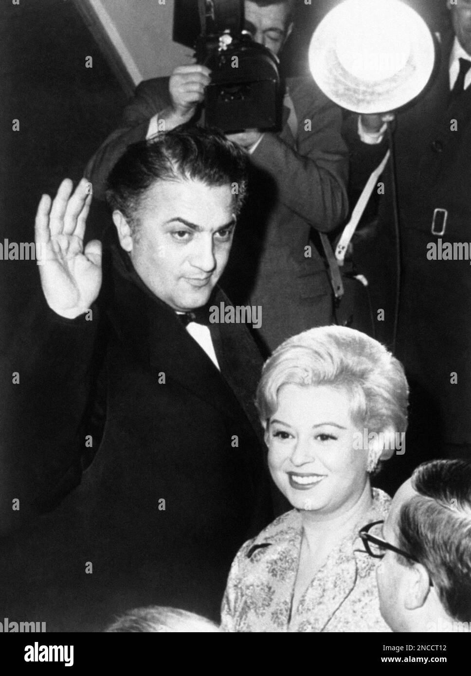 Italian movie director Federico Fellini waves to cheering crowd as he and his actress-wife Guilietta Masina, arrive at a fashionable Rome, Italy, movie theater on Feb. 13, 1963, to attend the premiere of Fellini’s latest film, “8 ½”, starring Italian actor Marcello Mastroianni and actress Claudia Cardinale. (AP Photo) Foto de stock