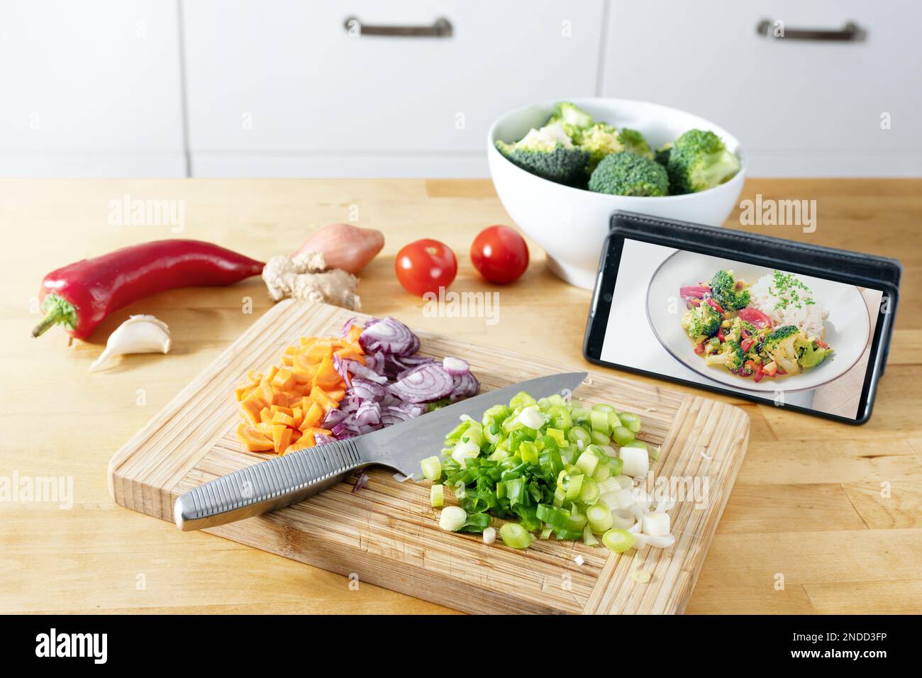 Cortar verduras en un tablero de cocina de madera, imagen de la receta terminada en el teléfono inteligente, cocinar mediante el uso de una aplicación en línea, preparación moderna de un HE Foto de stock