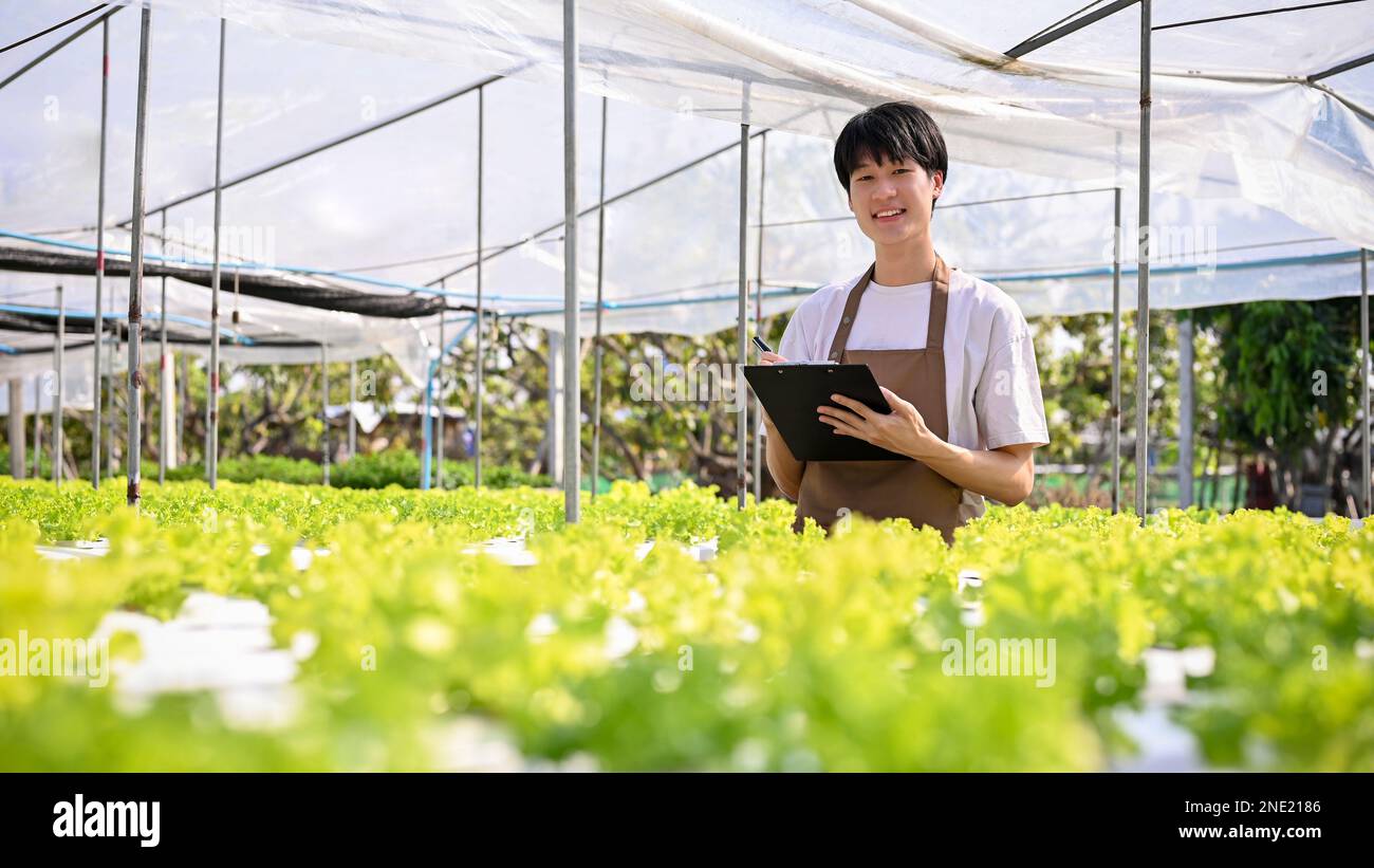 Feliz joven asiático hombre Pyme dueño de la granja orgánica que trabaja en el invernadero, registrando la calidad de las verduras de la ensalada en el portapapeles. Negocio de granja orgánica Foto de stock